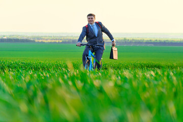 a businessman rides a bicycle with a backpack and a briefcase on a green grassy field, dressed in a business suit, beautiful nature in spring, freelance business concept