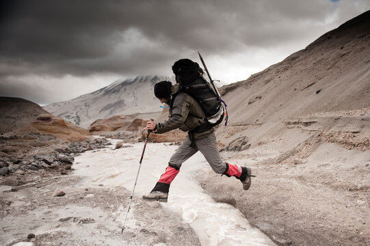 A mountaineer jumps across a glacial melt stream in the Valley of Ten Thousand Smokes Mount Griggs (2316m) can be seen in the background, Katmai National park, Alaska.
