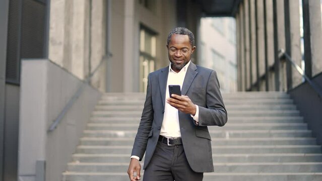 Smiling african american businessman checking text message while holding smartphone while walking outside Happy mature investor in suit is happy reading good news in front of stairs of office building