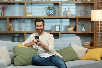 Young smiling man in a beige shirt sitting on sofa at home during quarantine. Holding the phone in his hands, he writes messages with his girlfriend, lover, friends, parents, relatives.