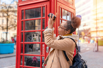 Fototapeta na wymiar Outdoor portrait of woman using camera against red phonebox in English city