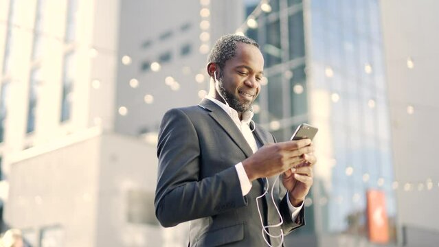 Smiling african american businessman with headphones using smartphone while standing outside. Mature confident male in formal suit reading text message, chatting online in front of office building