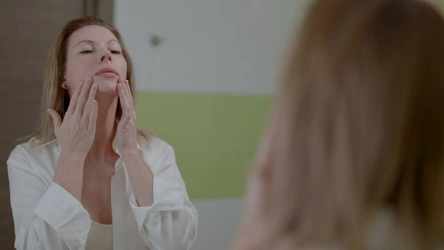 good-looking middle-aged woman looking her reflection in mirror in bathroom, massaging her neck