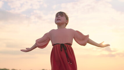 Fototapeta na wymiar Little girl prays against background of sky and sun. Religion and god, childhood dreams. Happy family. Child raised his hands to sky in park at sunset, true faith. Child plays in park against sky.