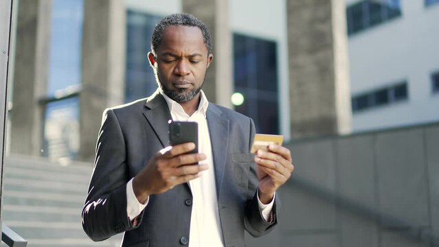 African american mature man entering credit card number on smartphone while standing outside in front of office building. Businessman in a suit is smiling because he made a successful purchase online