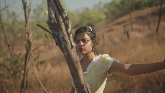 Dreamy 35mm film look of young, attractive Indian woman standing behind a tree in a sunny rural setting, looking flirtatiously at the camera before glancing downward, shallow depth of field.