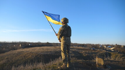 Portrait of soldier in military uniform lifted up flag of Ukraine in hill. Ukrainian army man holding waving flag. Victory against russian aggression. Invasion resistance concept. Slow motion