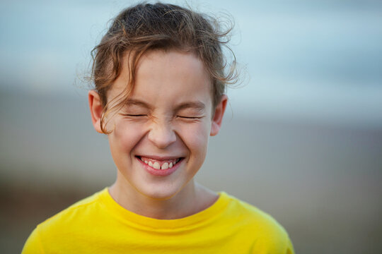 Close-up shot of a boy laughing with eyes closed tight. Happy child with curly hair in yellow t-shirt. Outdoor view against blurry background. Expecting a surprise