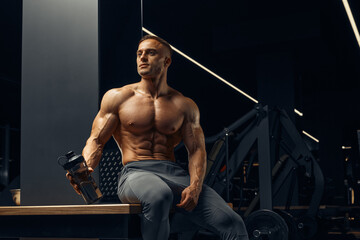 Fototapeta na wymiar Bodybuilder preparing to do exercises with dumbbells in gym. Bottle of protein in hand. Strength athlete sitting in gym on bench