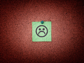 A green paper note with sad face on a cork board.