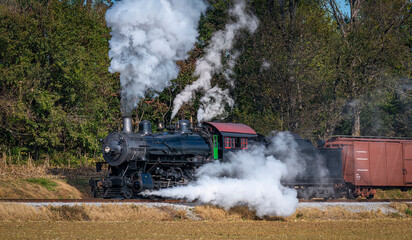 Fototapeta na wymiar Ronks, Pennsylvania, November 8, 2021 - A View of a Restored Steam Freight Train Blowing Smoke and Steam Traveling Along a Rural Countryside on a Sunny Day