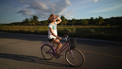 Side view of pretty mature senior woman biking into the sun wearing sunglasses and hat in a tropical setting.