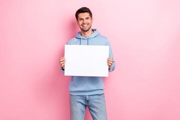 Photo of cheerful positive man wear blue hoodie holding white placard empty space isolated pink...
