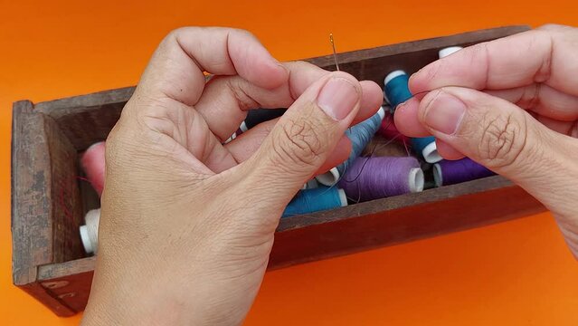 Footage of a human hand threading a needle with a white background and piles of colorful thread spools in a wooden box