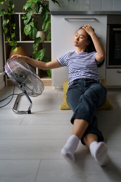 Relaxed young Asian woman lying on floor near working fan and enjoys coolness and fresh air. Carefree Chinese girl dressed in casual style is cooling off on summer day with electric climatic equipment