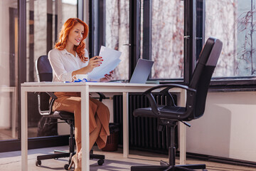 Happy smiling businesswoman in formal wear sitting at desk in modern office in front of laptop and...