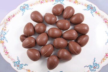 Group of Chocolate Easter eggs on a white plate with traditional decorations of bows and flowers and a pale background.