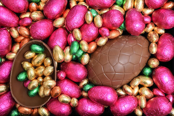 Pile or group of multi colored and different sizes of colourful foil wrapped chocolate easter eggs in pink, red, gold and lime green with two halves of a large brown milk chocolate egg in the middle.