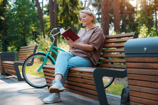 Dreamy modern old lady relaxing in city park. Pensive senior grey haired woman in casual sitting wooden bench outdoors.