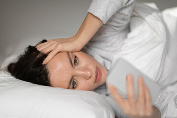 Sad unhappy young european woman lies on white bed, wakes up, reads message on smartphone in bedroom