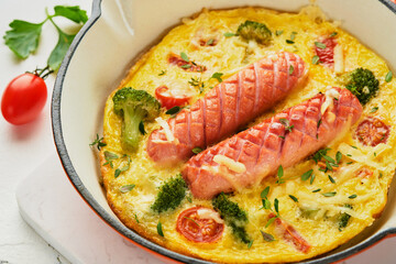Omelette or frittata with two sausages, melted cheese, cherry tomatoes, broccoli and parsley on red skillet or iron pan on white old concrete background. Healthy breakfast food concept. Top view.