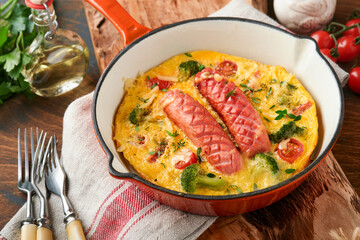 Omelette or frittata with two sausages, melted cheese, cherry tomatoes, broccoli and parsley on red skillet or iron pan on old wooden rustic table background. Healthy breakfast food concept. Top view