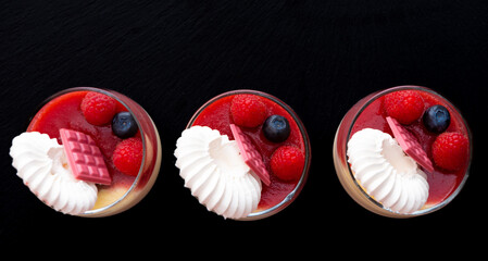 three glasses with red raspberry jelly and white panna cotta on black background. Image from above....