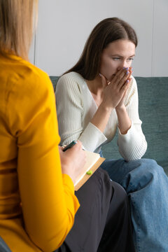 Young Sad Woman Is Sitting On The Couch, Holding Her Hands To Her Face At A Psychotherapist's Appointment. Psychologist With A Client. Photo From The Psychologist's Back. Vertical Photo