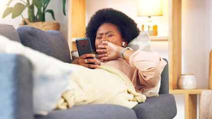 Bored and tired woman lying on a couch typing on her phone at home. A young African female relaxing...