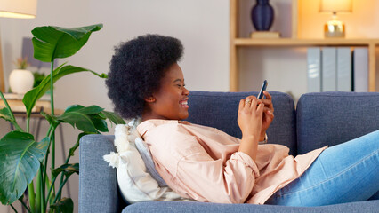 A laughing young woman texting on a phone at home. Cheerful female chatting to her friends on social media, browsing online and watching funny internet memes while relaxing on a sofa - obrazy, fototapety, plakaty