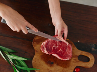 Woman with knife in hand cutting fresh steak meat for roasting kitchen with salt pepper other spices on table, red cherry tomatoes and herbs, preparing dinner in home kitchen. wooden table, top view.