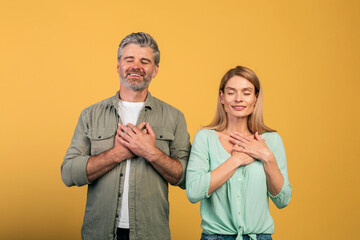 Thank you. Grateful middle aged couple with closed eyes keeping both hands on chest, expressing...