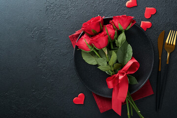 Valentines day romantic table setting. Empty closeup of red roses, wine, candles, dinner black plate, knife, fork and decorative silk hearts on black background. Holiday concept. Copy space. Top view