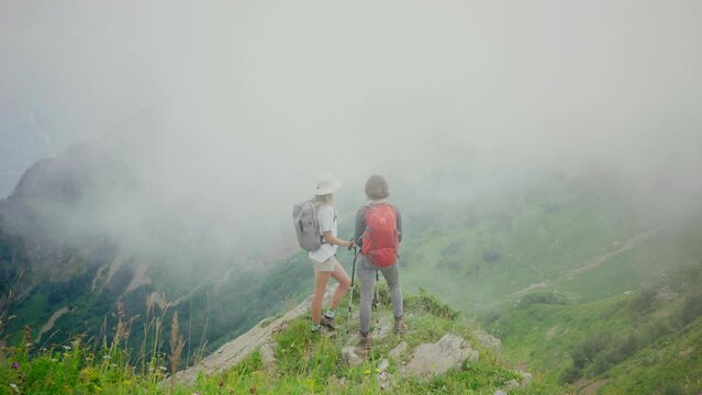 Two women with backpacks and trekking poles hiking in mountains amidst meadows 