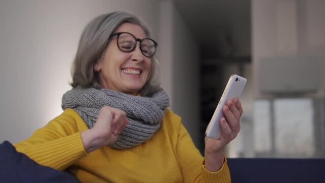 Extremely happy excited woman listening to favourite music on smartphone application and dancing, sitting on sofa, having fun enjoying performance online, excitement