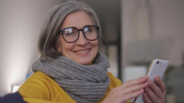 Happy self-confident woman with gray hair in stylish eyeglasses holding smartphone in hands and smiling, looking at camera, working from home, receiving additional income, advertisement