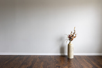 Tall, clay vase with dry plants arrangement next to an empty white wall in a room with laminated floor.