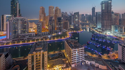 Dubai Marina with several boats and yachts parked in harbor and skyscrapers around canal aerial night to day .