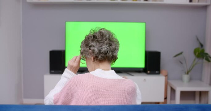 Senior lady sits on a couch and turns on TV with greenscreen, chroma key, 4k