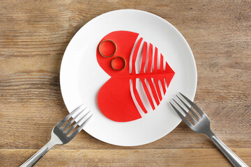 Divorce concept. Plate with paper cutout of heart, wedding rings and forks on wooden table, flat lay