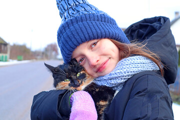        A ten-year-old girl in a blue hat with a multi-colored kitten on a village street, close-up