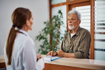 Senior man talking to his doctor while filling medical documents in hospital.