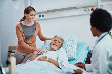 Happy woman and her hospitalized senior father talking to black female doctor at medical clinic.