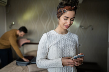 couple young beautiful woman and man in hotel room wife or girlfriend use mobile phone for texting sens sms messages while husband or boyfriend is unpacking or packing baggage on the bed copy space © Miljan Živković