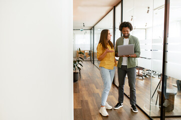 Two colleagues discussion project standing together in te hallway, man and woman looking at laptop screen and solving business tasks in relaxed office atmosphere, talking in order to reach a decision