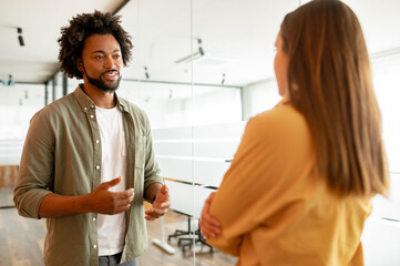 Two coworkers chatting during coffee break standing in glass hall of office building. Curly...