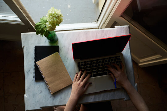 Unrecognizable Girl Working On Laptop At Marble Desk. Overhead Of Unrecognizable Woman Typing On Laptop At Marble Table With Flowers And Notebooks At Opened Door.