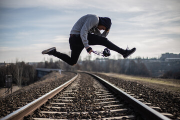 man jumping over train tracks. man with a camera. man wearing a hat. urban photography concept