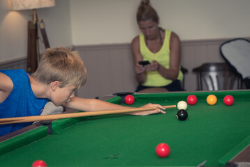 A boy plays billiard or pool in club. Young Kid learns to play snooker. Boy with billiard cue strikes the ball on table. Active Leisure, sport, hobby concept