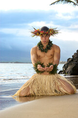Male Hula Dance poses on the beach as he kneels in the sand in a giving gesture. 